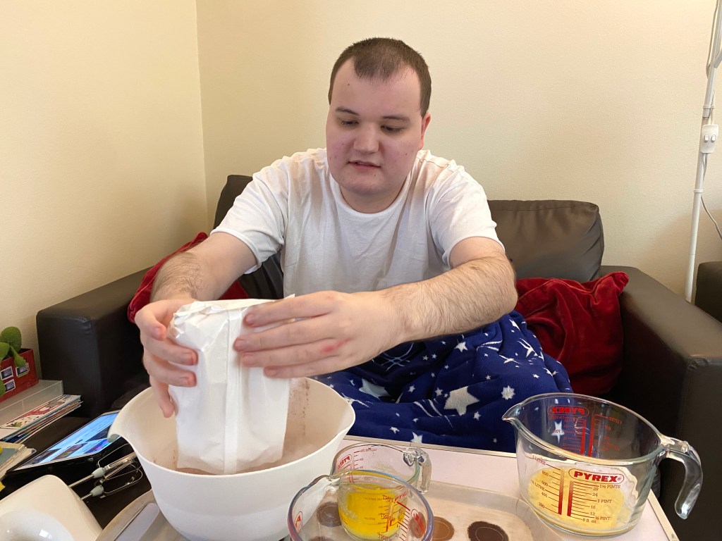 A person in a white t-shirt is pouring flour from a bag into a mixing bowl while seated on a couch, wearing blue patterned pajamas. Various measured ingredients, including eggs and melted butter, are present on the table.