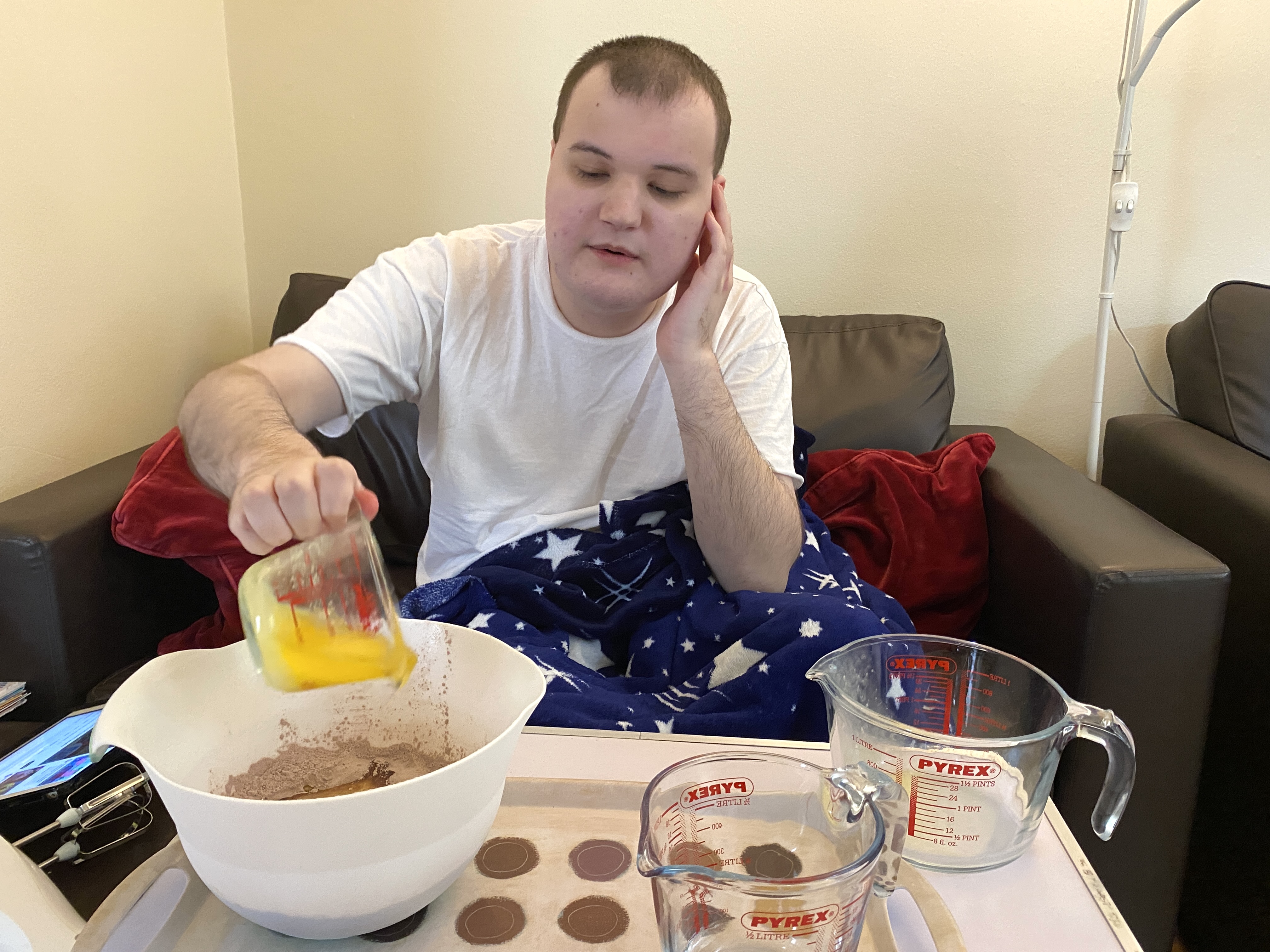 A person sitting on a couch, wearing a white t-shirt and a blue blanket with stars, is pouring an egg from a glass measuring cup into a mixing bowl filled with brown ingredients.