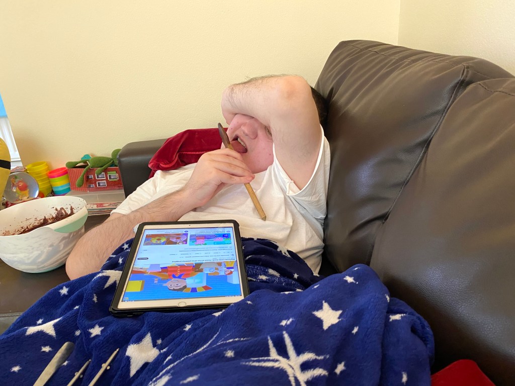 A person lounging on a couch covered with a star-patterned blanket, holding a pencil and watching a tablet screen displaying a colorful children's cartoon, with a bowl of chocolate mixture nearby.