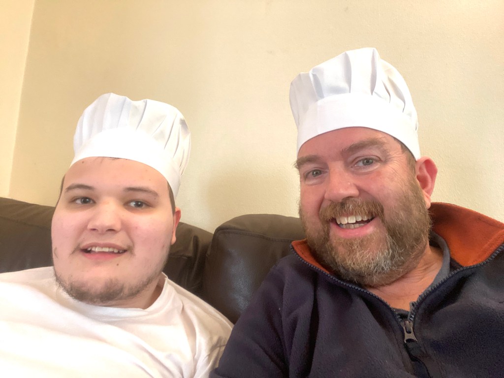 Two smiling individuals, one young and one adult, wearing white chef hats while sitting on a couch.